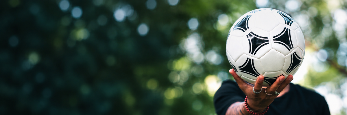 Una persona sostiene un balón de fútbol frente a un fondo natural desenfocado.